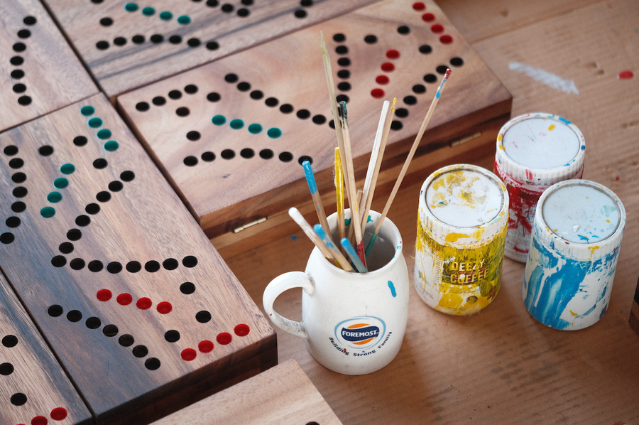 Deux plateaux de jeu de Tock peints à la main, pots de peinture et pinceaux posés sur l'établi