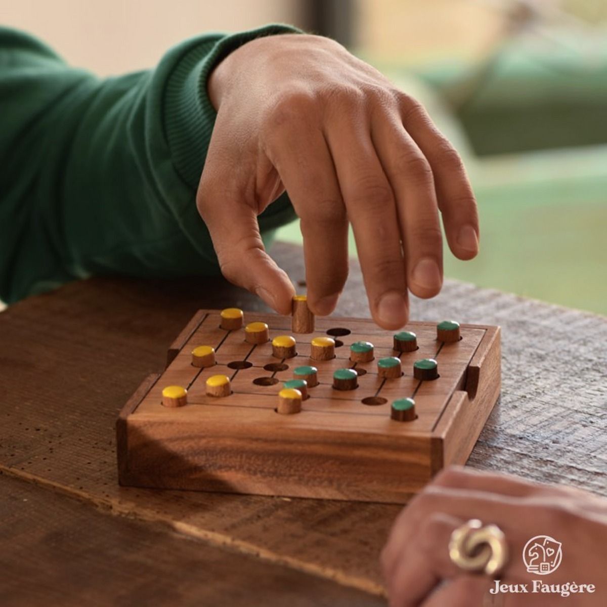 Marelle en bois Jeux Faugère, jeu du Moulin : une main pose un pion jaune sur le plateau en albizia, face aux pions verts