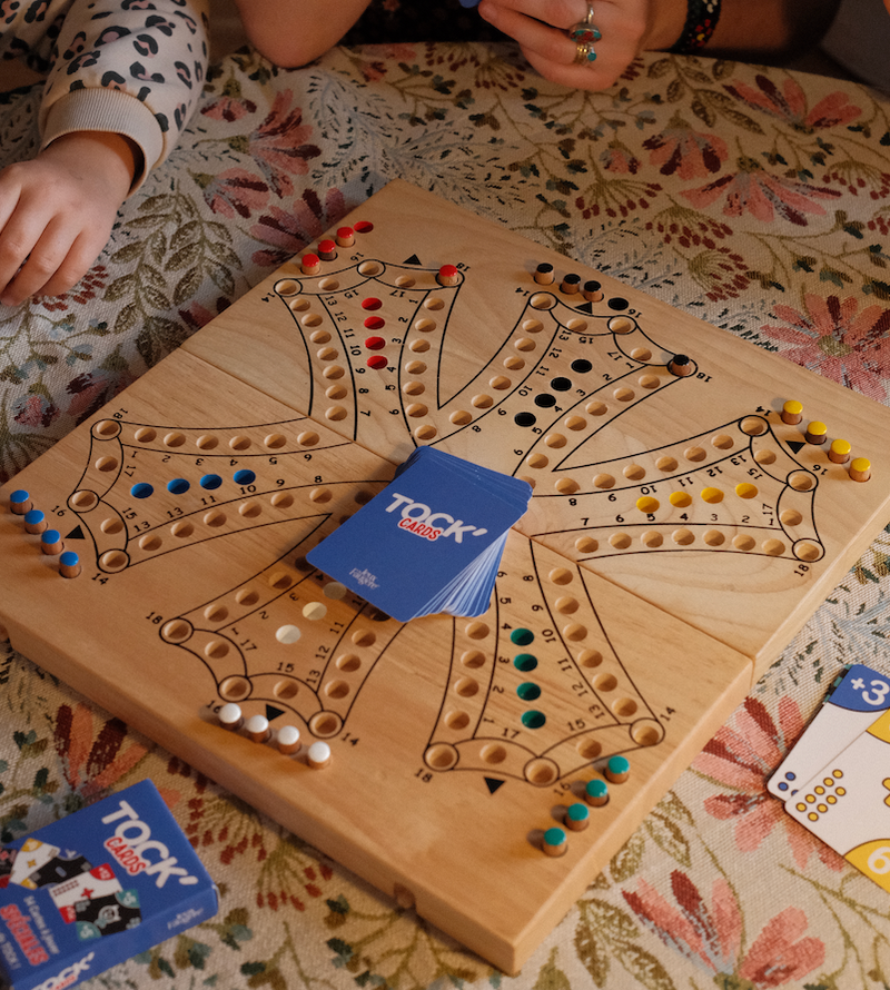 Grand plateau de jeu de Tock en bois hévéa clair pour 6 joueurs, avec le jeu de cartes Tock'CARDS Jeux Faugère au centre et les pions des six couleurs en place