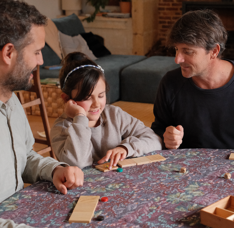 Une fillette et deux adultes jouent à un jeu de mémoire en bois autour de la table familiale