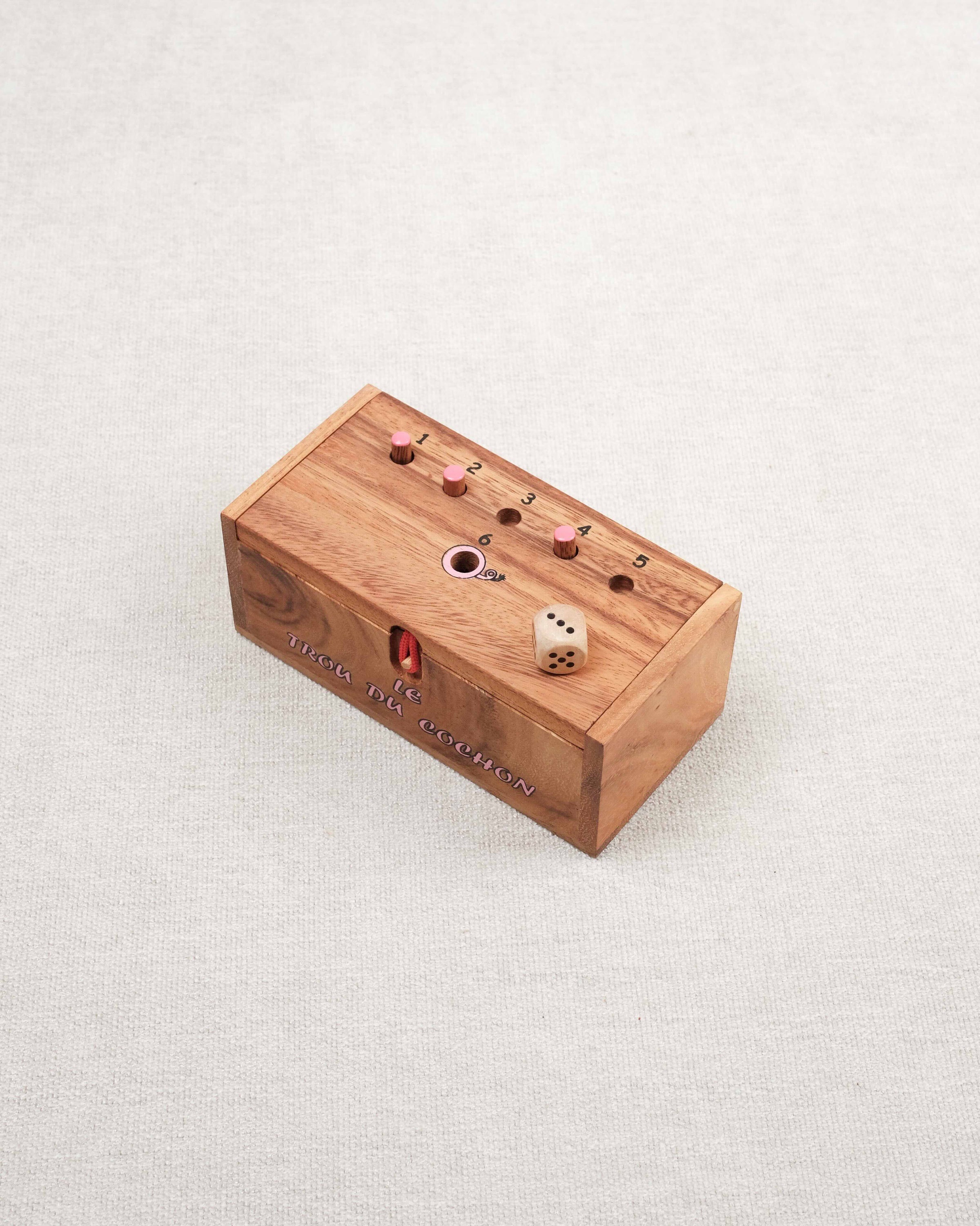 Coffret du jeu "Le trou du cochon" avec dé en bois et pions roses sur le dessus