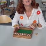 Une enseignante présente le jeu Ferme la boîte en bois sur une table de sa classe, avec les clapets numérotés et les dés