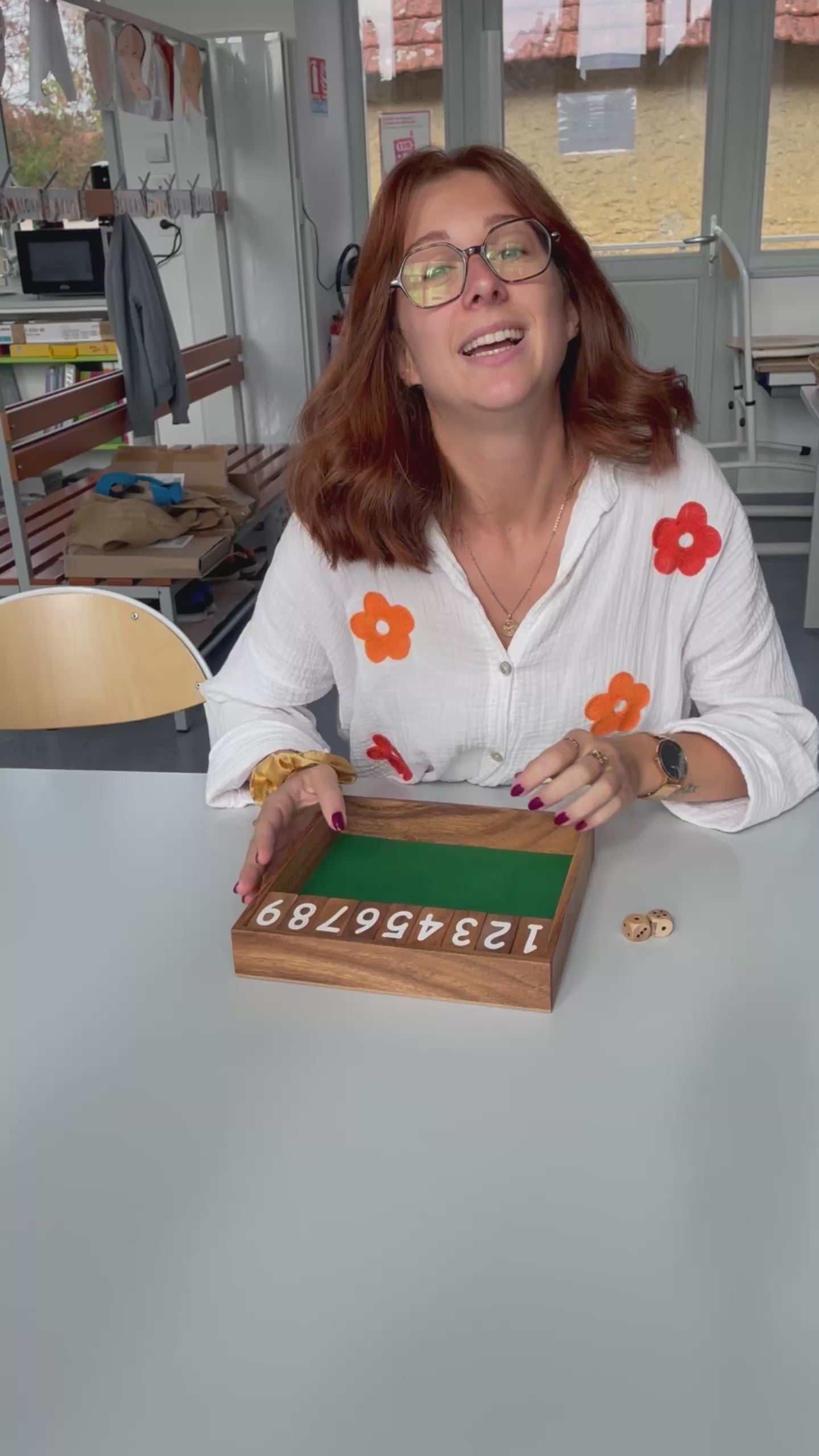 Une enseignante présente le jeu Ferme la boîte en bois sur une table de sa classe, avec les clapets numérotés et les dés
